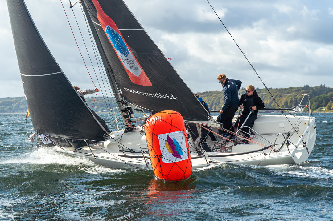DCC 2021; Segelboot Typ Corby mit schwarzen Segeln. Auf dem Großsegel ist eine orange Manöverschluck Falsche z sehen. Man sieht auf dem Bild ein Mann an der PInne und einer am Großfall. Vor dem Boot seiht man eine aufgeblasene Regattatonne mit dem Logo vom Deutschen Company Cup.